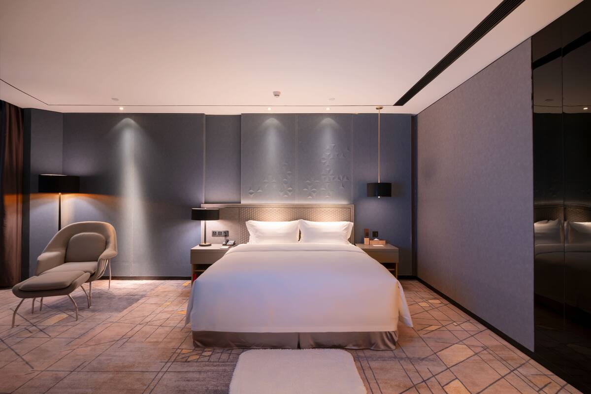 A modern hotel room with a large bed, a bedside table, a chair, and a large mirror.