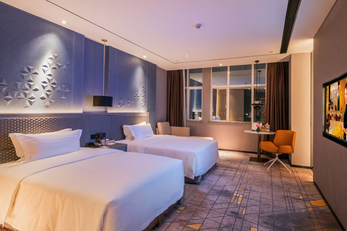 A hotel room with two beds, a desk, a chair, a window with curtains, and a wall with a geometric design.