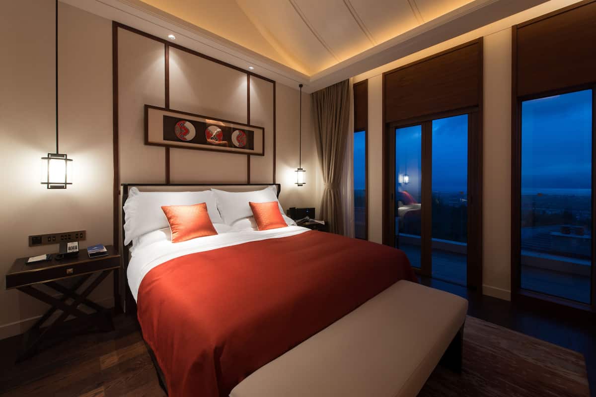 Une chambre d’hôtel avec un grand lit, des tables de chevet, une petite table, un tableau encadré au mur et une vue sur la ville par les fenêtres.