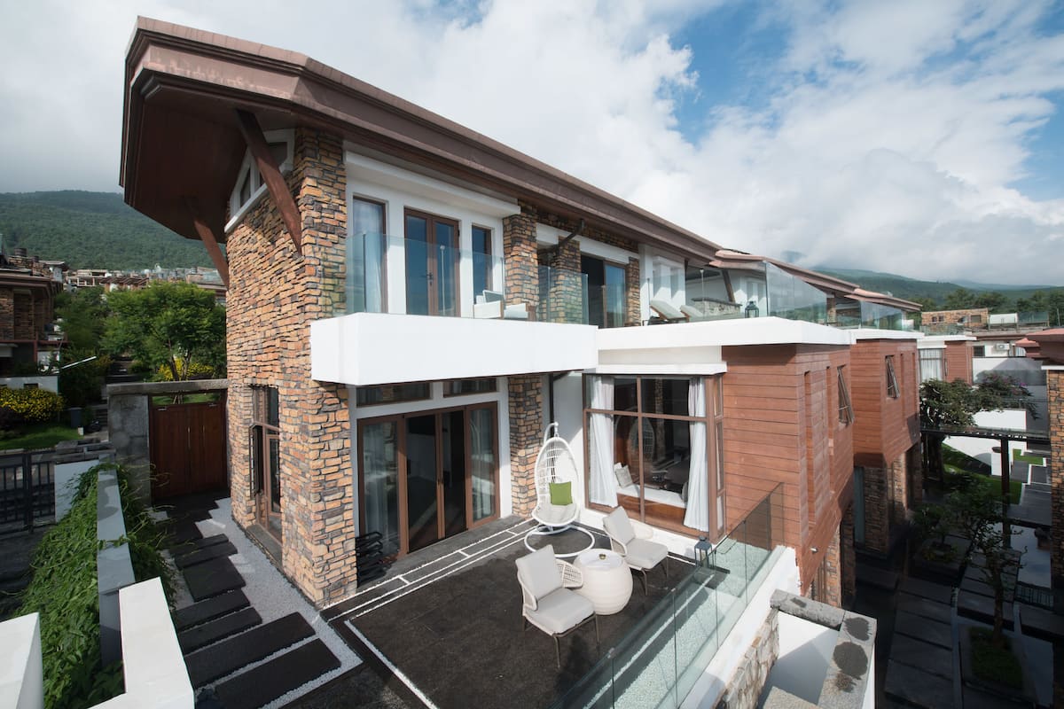 Une maison moderne à deux étages, dotée d’un balcon en verre, d’une façade en pierre et offrant une vue sur le paysage environnant.