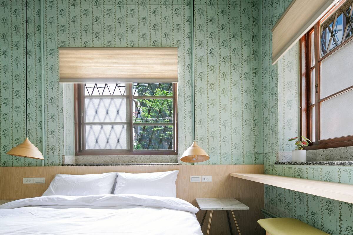 A bedroom with a bed, two bedside lamps, a window with a view of trees, and a small table.