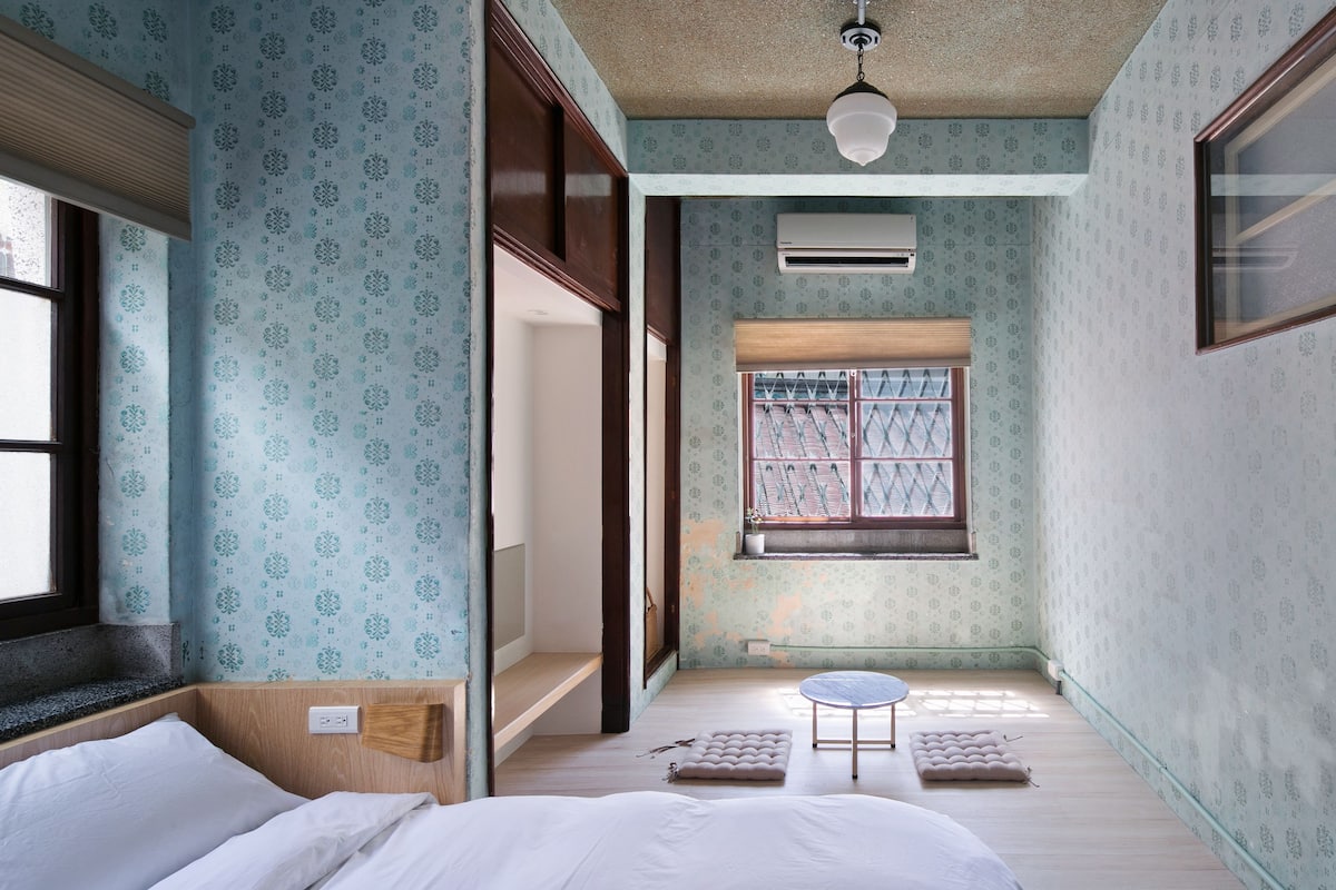 A compact room with patterned wallpaper, a bed, a small round table, a window with a view, and a TV on the right wall.