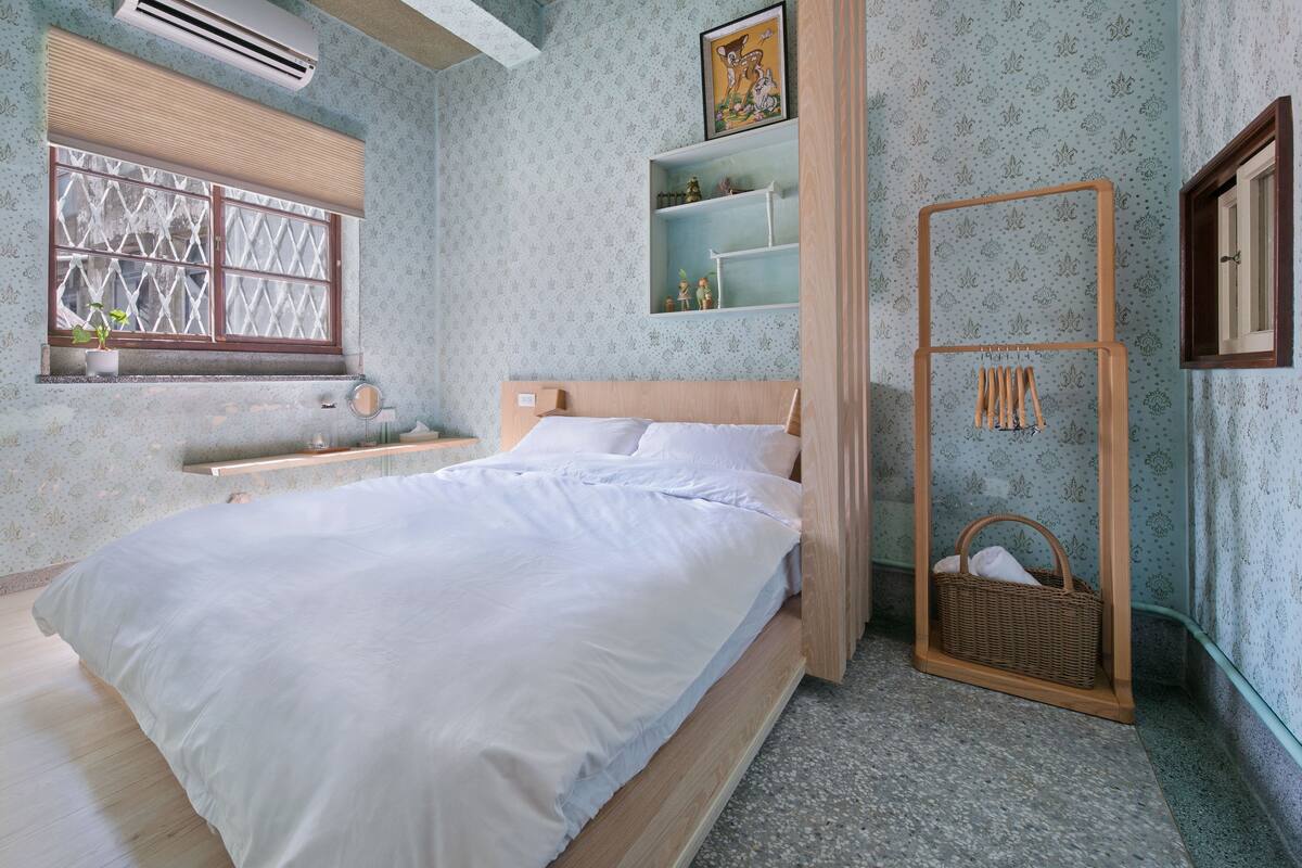 A bedroom with a bed, a window with blinds, a shelf with decorative items, and a wall-mounted rack with hanging items.