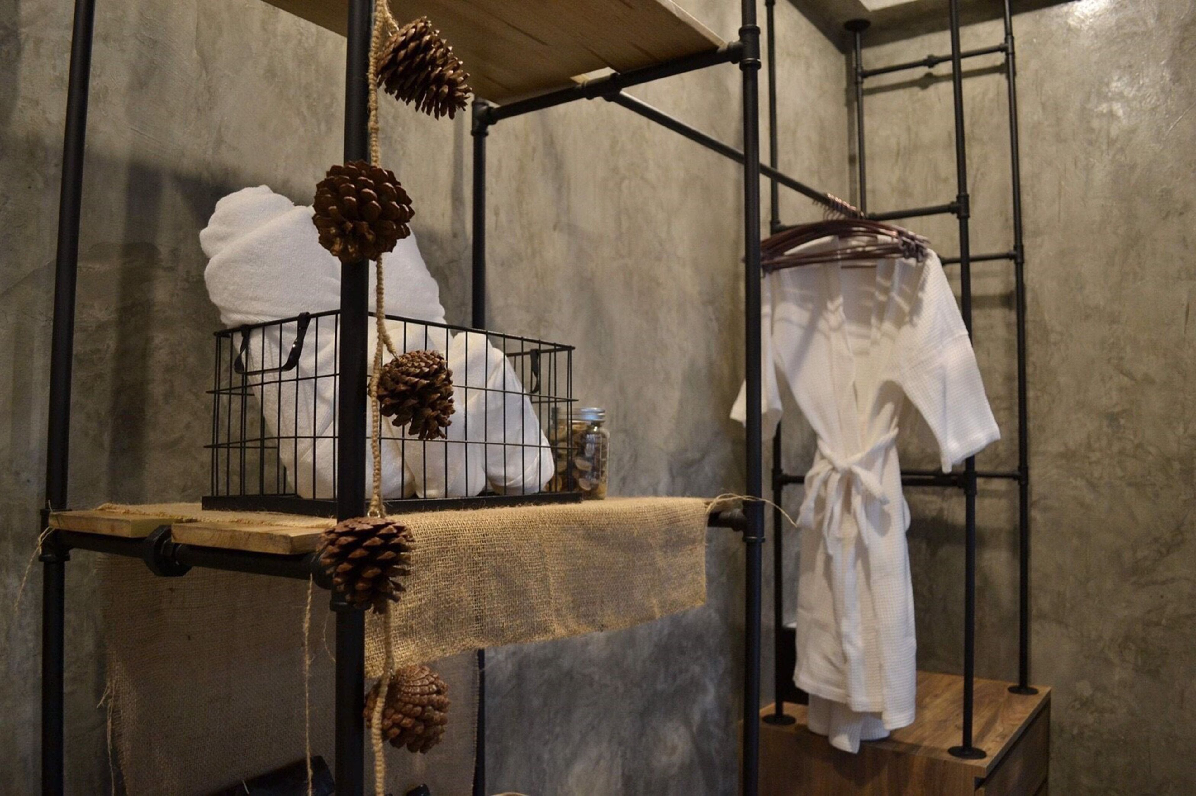 A rustic bathroom with a metal rack holding towels and a robe, a wooden shelf with pine cones, and a concrete wall.