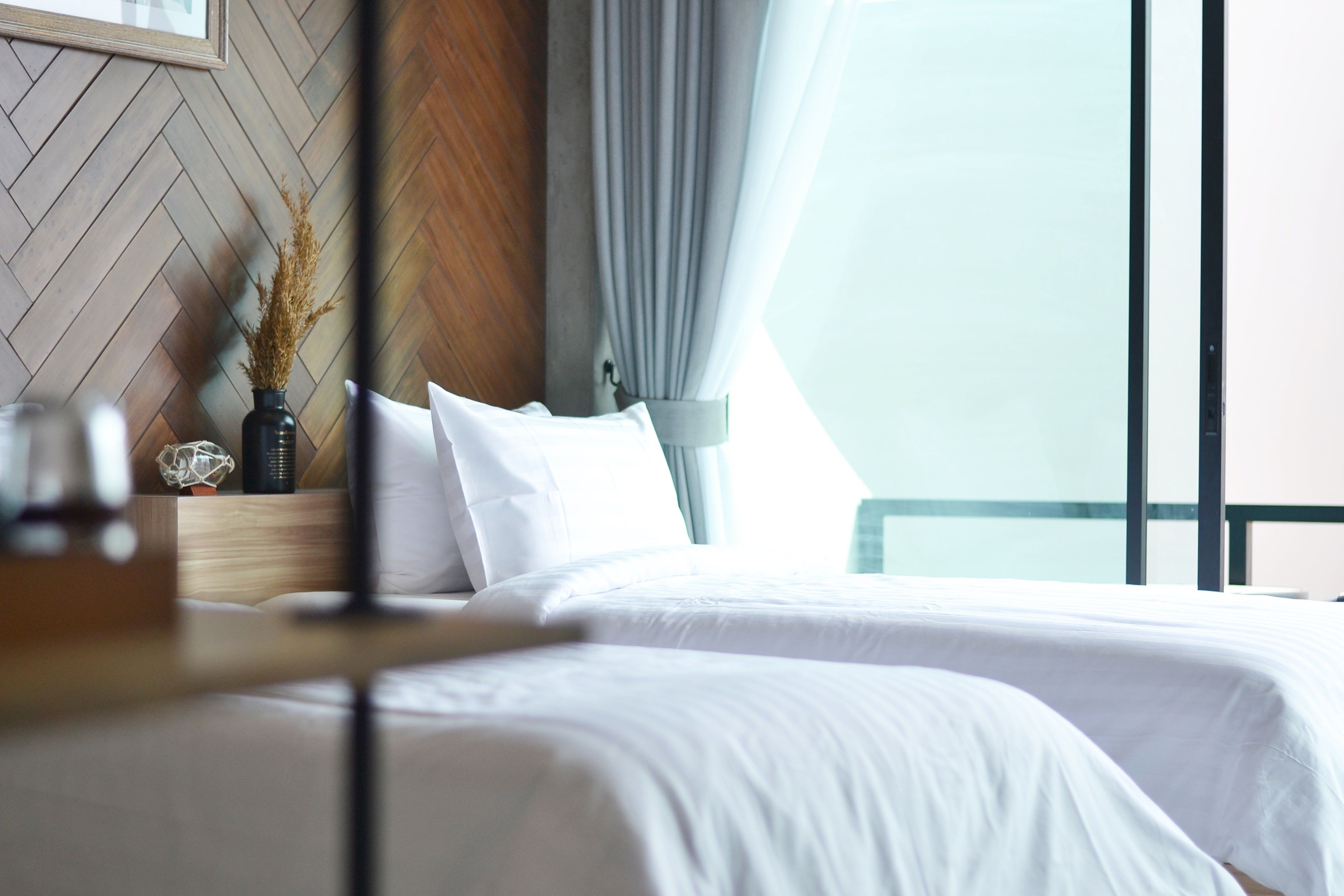 A neatly made bed with white linens, a wooden headboard, and a vase with dried plants.