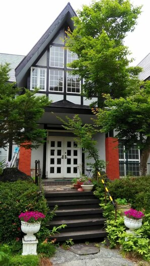 A two-story house with a steeply pitched roof, a front door with glass panels, and a small front porch with steps leading to the entrance. - Pension Galaxy (Katashina)