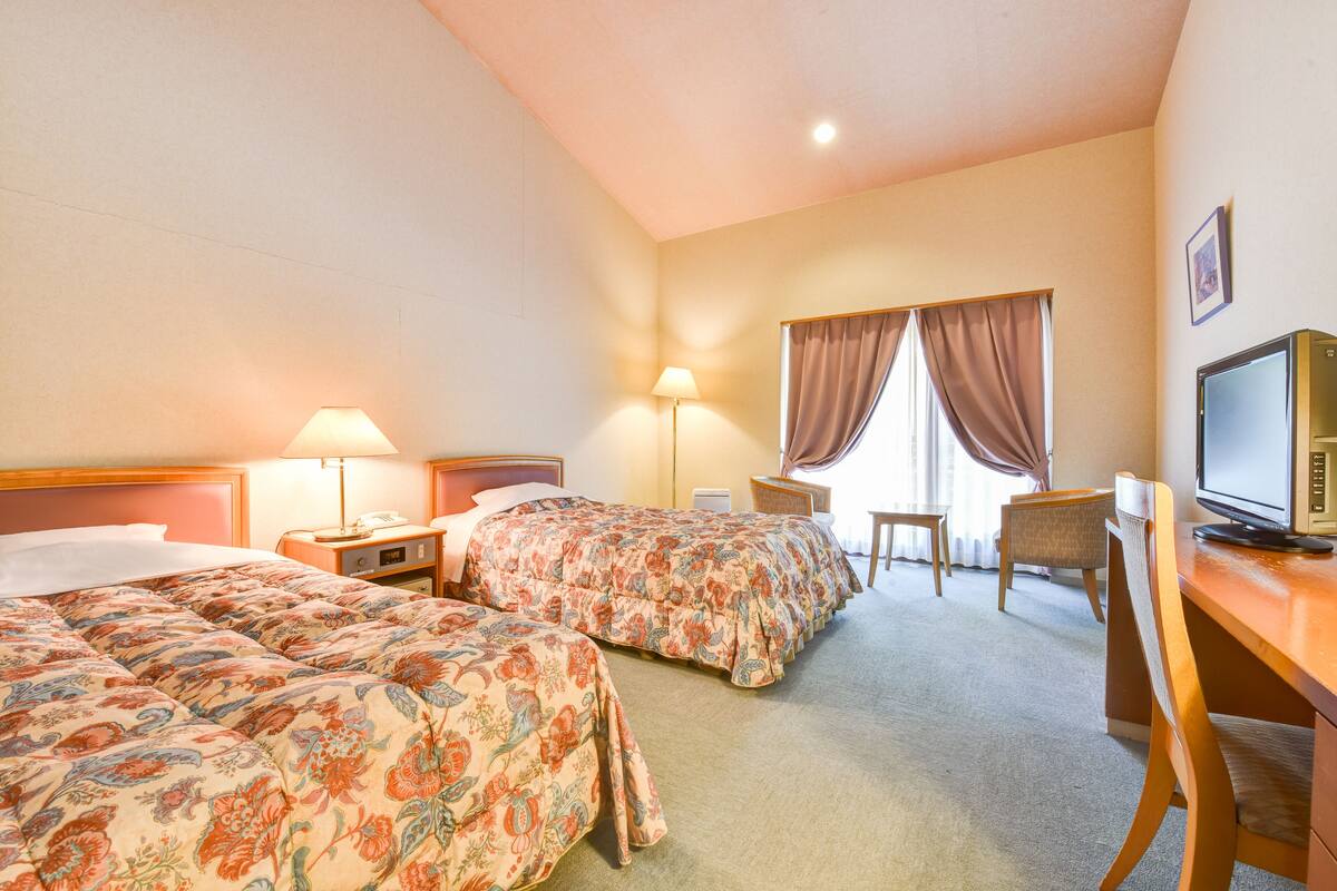 Quarto de hotel com duas camas, uma escrivaninha, uma cadeira, uma televisão e um abajur.