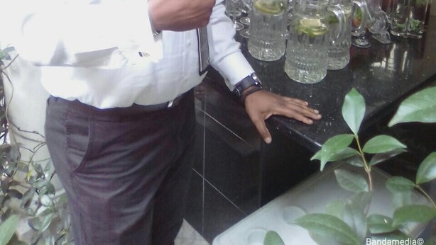 Un hombre con camisa y corbata de blanco, parado junto a una barra con varios vasos y botellas.