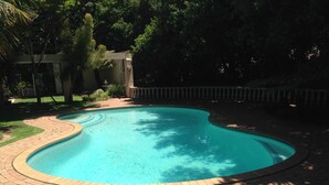 A swimming pool with a curved edge, surrounded by a brick walkway and a grassy area. - Noraclox Riverclub (Sandton)