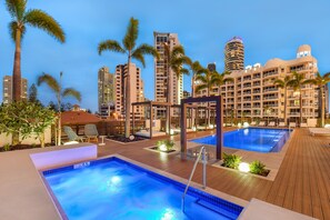 Un área de piscina en la azotea con dos piscinas, sillas de descanso y palmeras.
