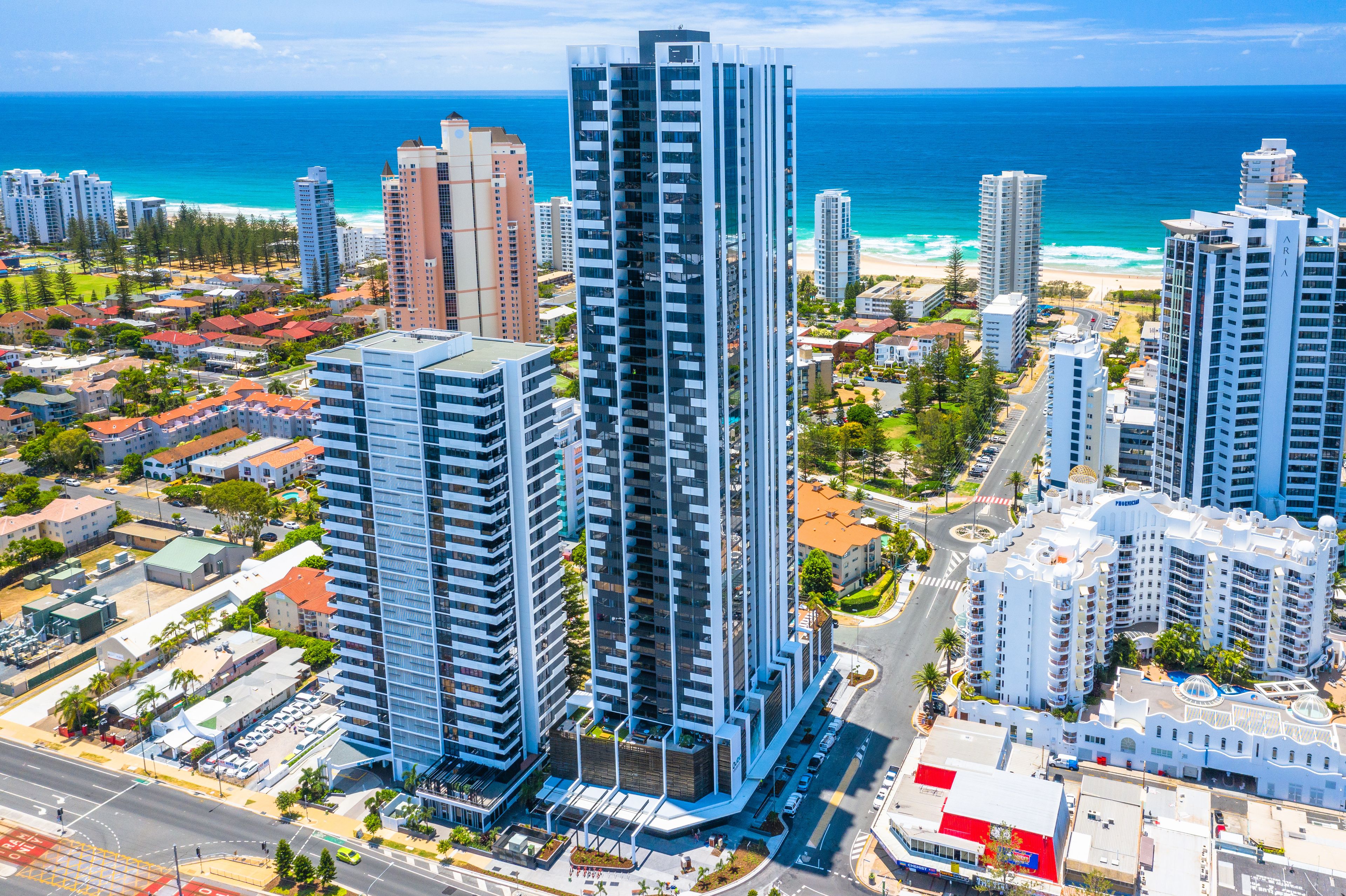 Un edificio de gran altura con diseño moderno, rodeado de otros edificios residenciales y una playa al fondo.
