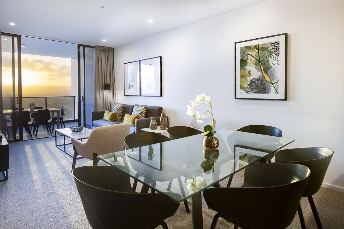 Ein modernes Wohnzimmer mit einem Glastisch, schwarzen Stühlen, einer Couch mit gemusterten Kissen und einem Balkon mit Blick.