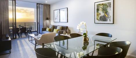 A modern living room with a glass dining table, black chairs, a sofa with patterned cushions, and a balcony with a view.