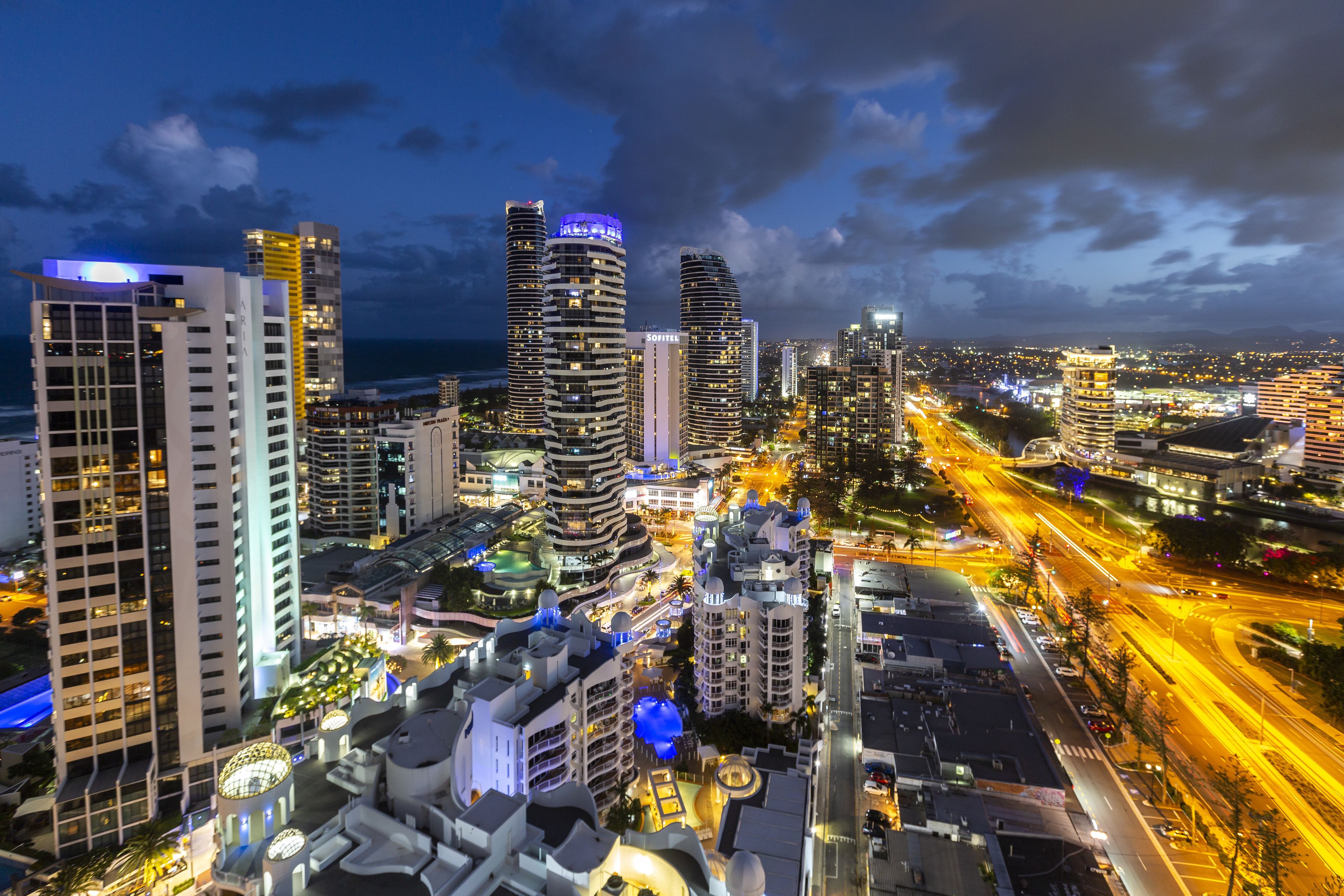 A cityscape with modern high-rise buildings, a well-lit street, and a view of the ocean.