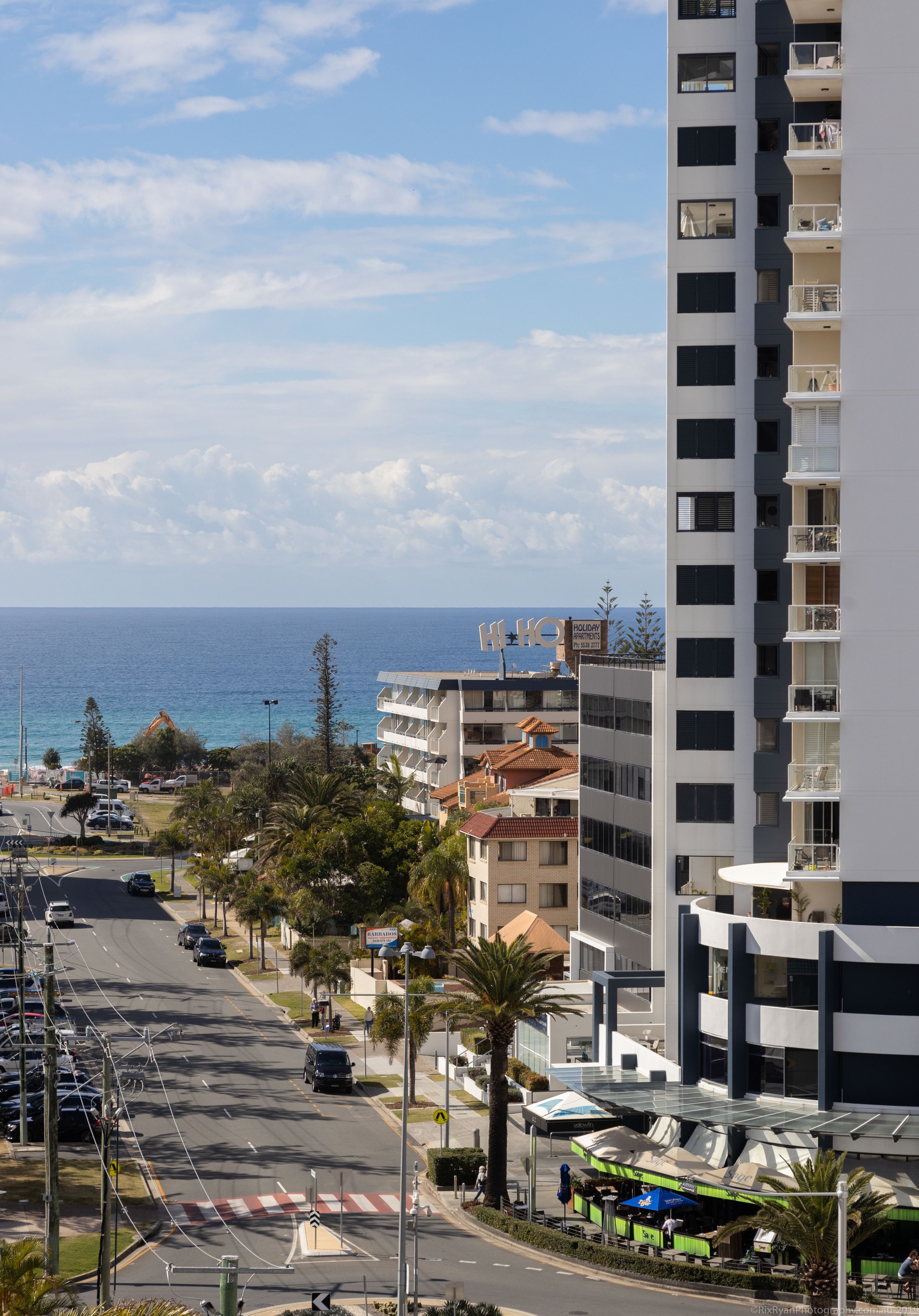 A coastal urban area with a modern hotel, a clear view of the sea, and a wide street with parked cars.