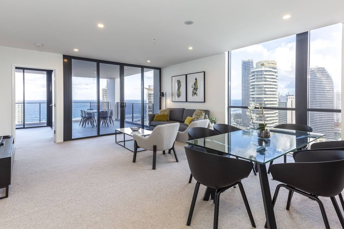 Ein geräumiger Wohnbereich mit Glastisch, schwarzen Stühlen, einer grauen Couch und einem Balkon mit Blick auf das Meer und die Skyline der Stadt.