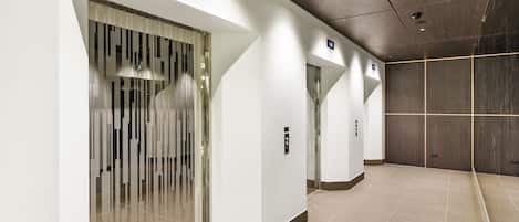 Un área de ascensor moderna con puertas de vidrio, una pantalla digital y suelo de baldosas.
