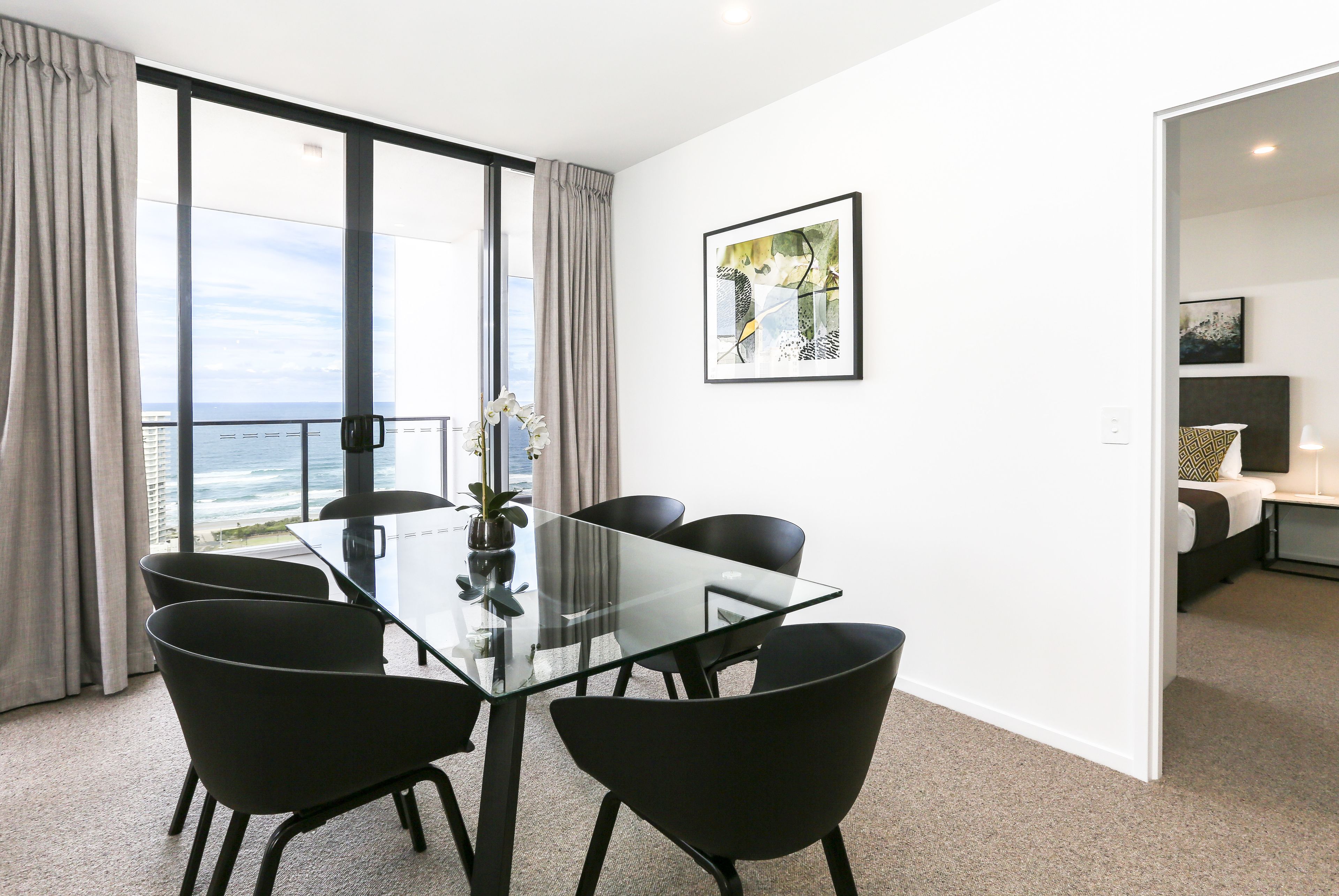 A modern room with a glass table and black chairs, a large window with a view of the ocean, and a framed picture on the wall.