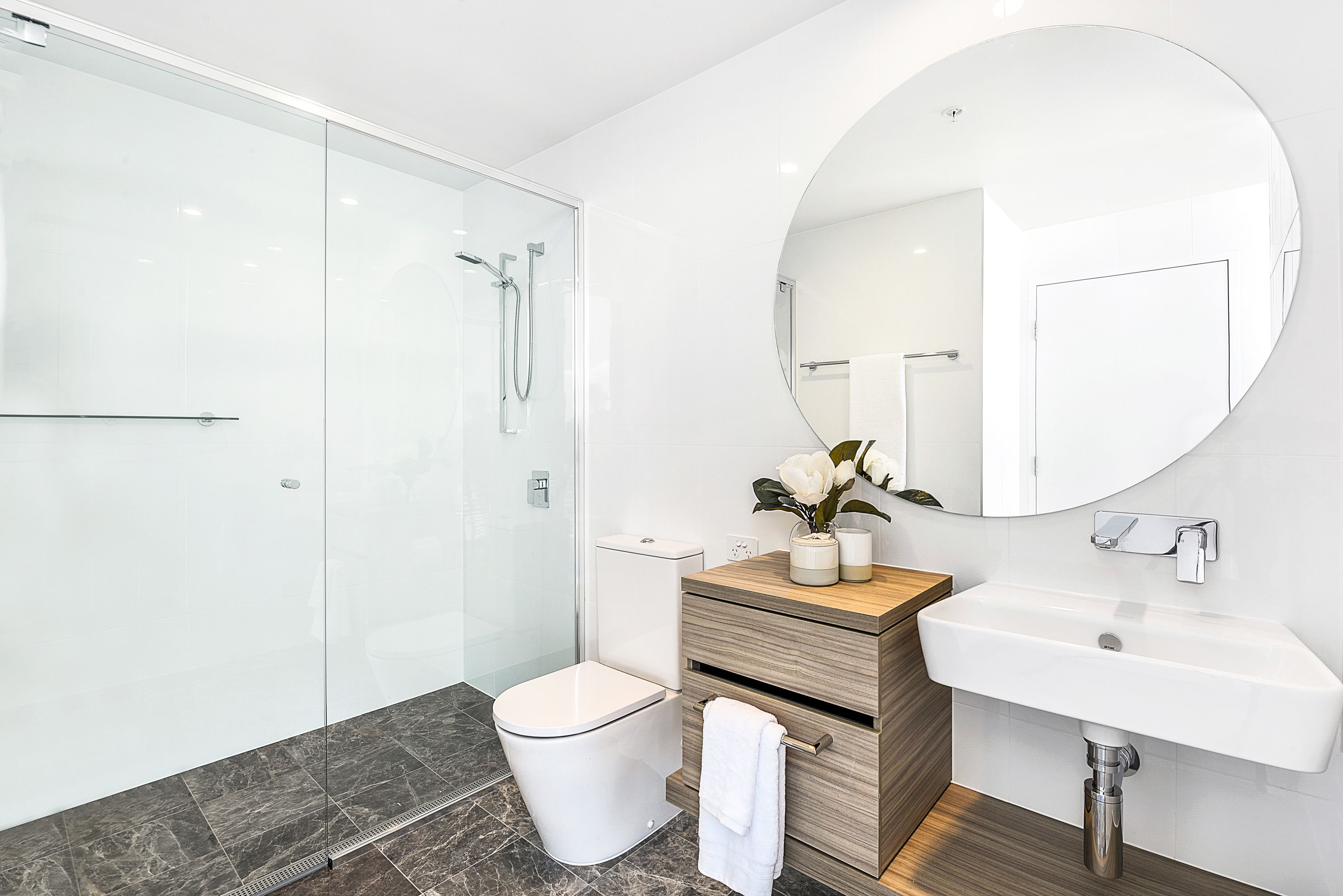 A modern bathroom with a glass shower enclosure, a white toilet, a wooden vanity with a sink, and a large mirror.