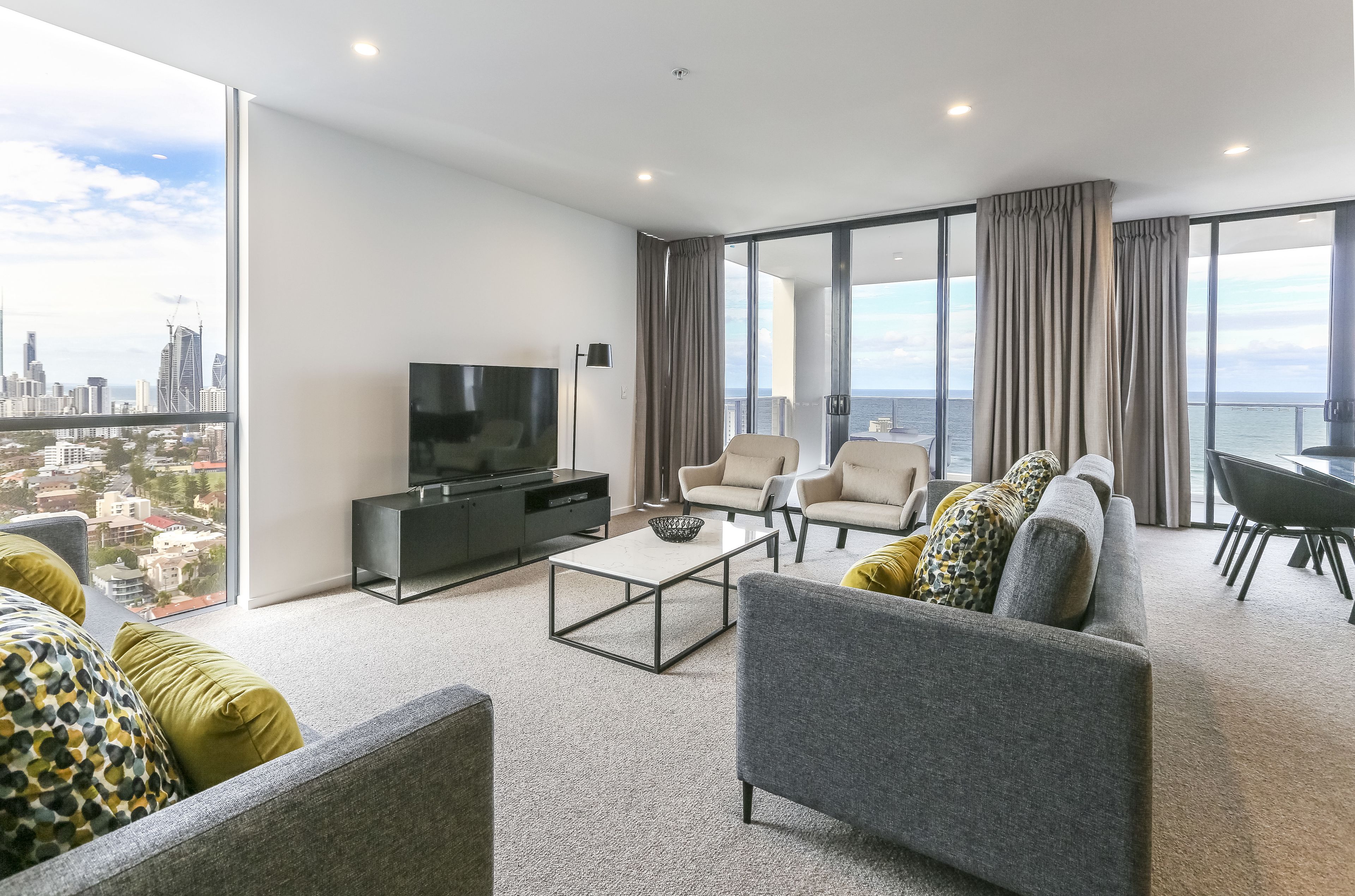 A modern living room with a large TV, comfortable seating, and a view of the city and sea.