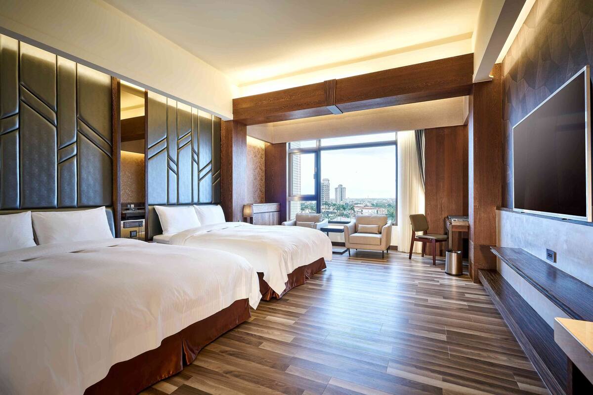 A modern hotel room with two large beds, a seating area with a view, and a large TV on the wall.