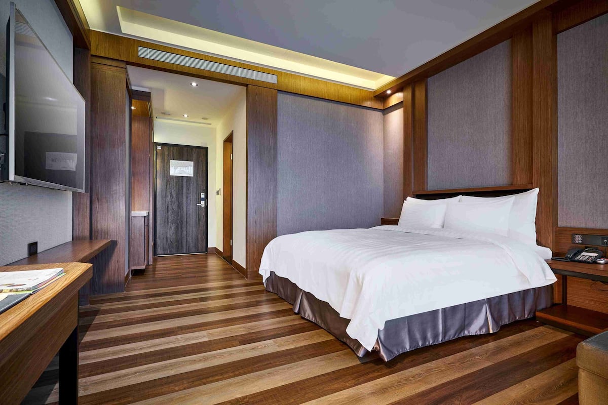 A hotel room with a large bed, wooden flooring, a flat-screen TV, a desk, and a door leading to another room.