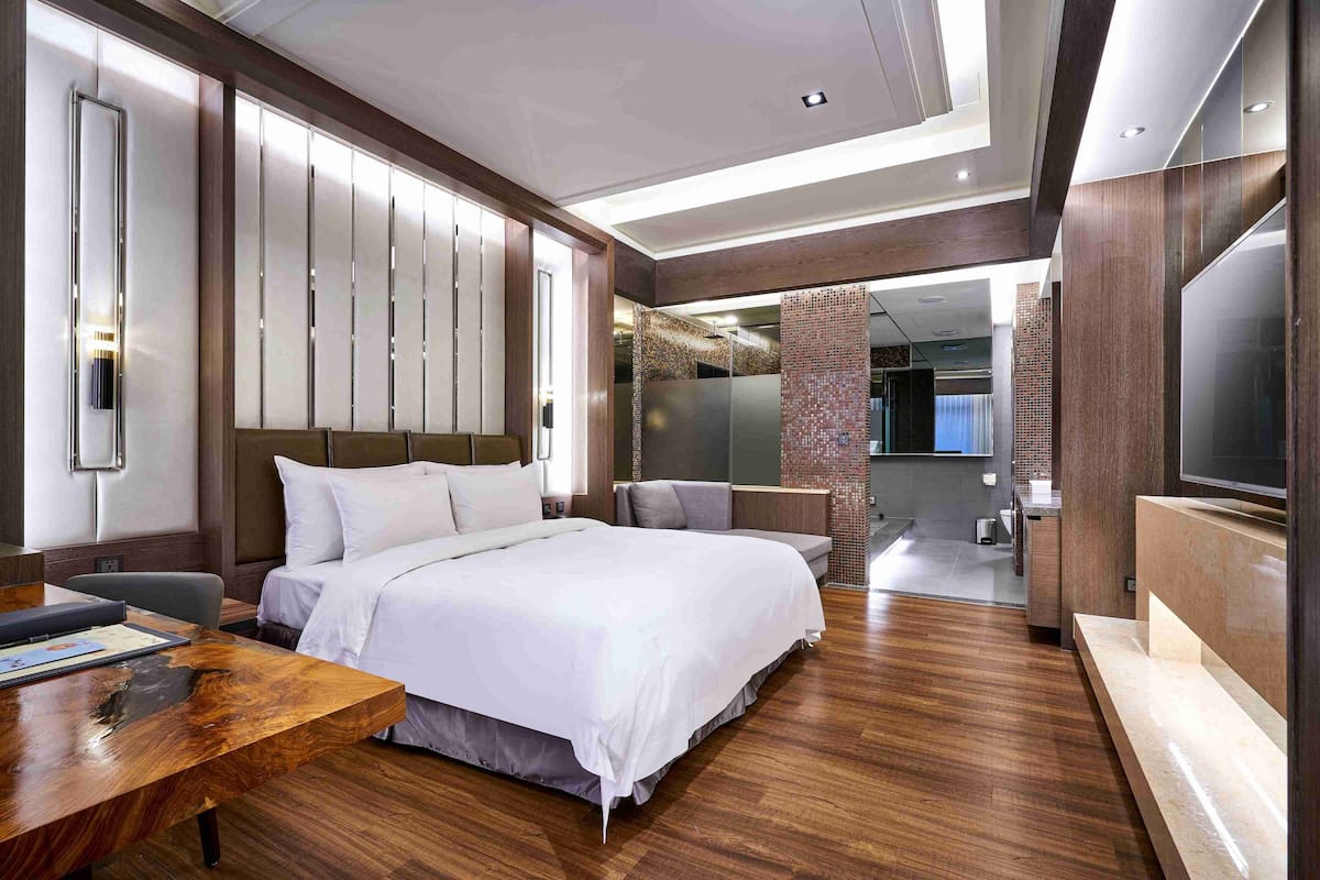 A modern hotel room with a large bed, a wooden desk, a TV, and a bathroom visible through a glass partition.