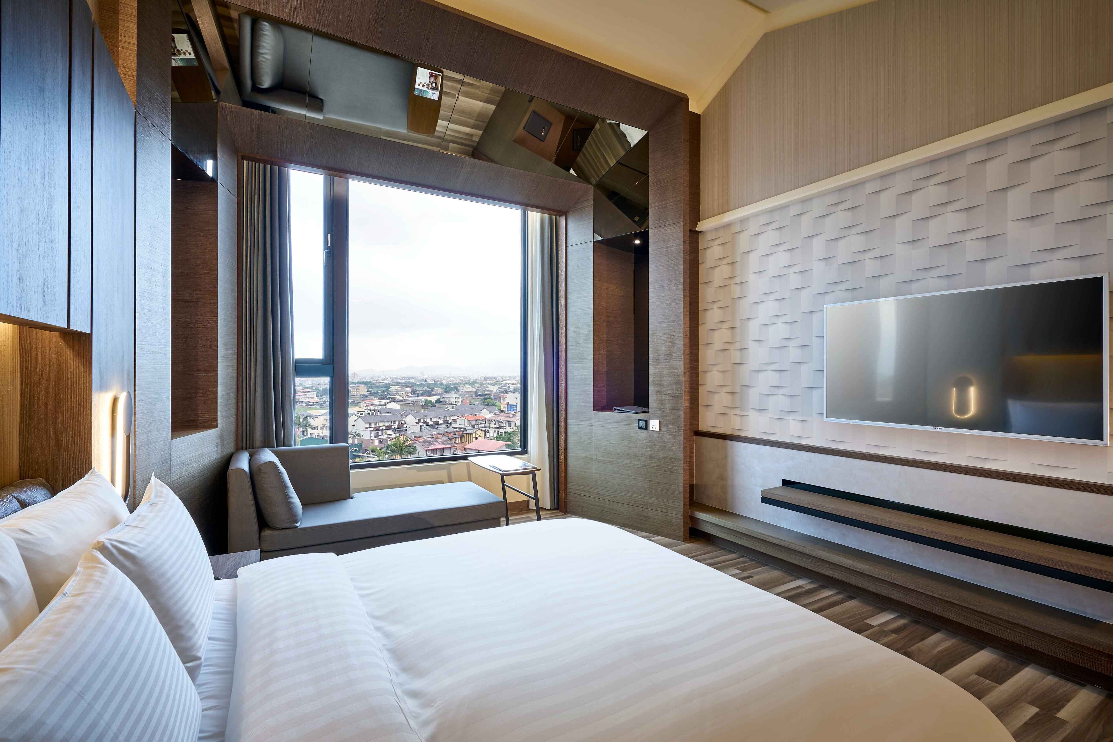 A modern hotel room with a large bed, a TV mounted on the wall, and a view of the city through a large window.