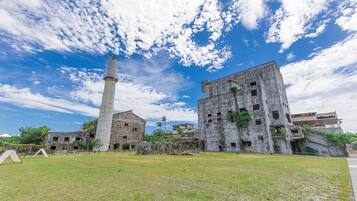 An abandoned building complex with a tall chimney, overgrown vegetation, and a grassy area in front.