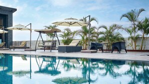 A pool area with lounge chairs and umbrellas, a modern building with large windows, and palm trees in the background. - So Fun Hotel (Wujie)