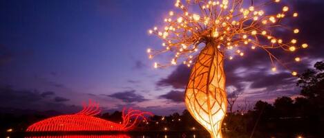 A night scene with illuminated structures, including a tree-like sculpture with glowing lights and a red-lit bridge.