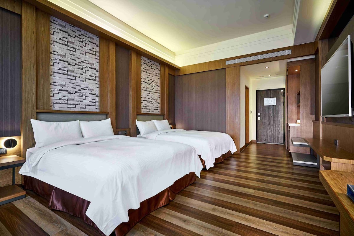A hotel room with two beds, wooden flooring, a stone accent wall, and a TV on the right.