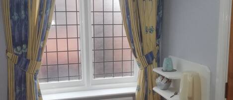 Una habitación con ventana, cortinas, un radiador, hervidor, un tazón y un cubo de basura.