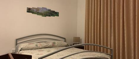 A bedroom with a metal-framed bed, a wooden chair, a wall-mounted lamp, and a window with beige curtains.