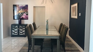 Een eethoek met een tafel met glazen blad, donkere gestoffeerde stoelen, een moderne kroonluchter, een stadslandschap schilderij en een ingelijste foto aan de muur.