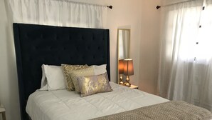 Een keurig opgemaakt bed met een donker hoofdbord, witte beddengoed en decoratieve kussens. Er staat een bedlampje en er hangt een spiegel aan de muur.