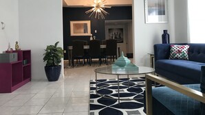 Een moderne woonkamer met een blauwe bank, een salontafel, een kamerbreed tapijt, een eetgedeelte met een kroonluchter, en een donkere wand met een ingelijst kunstwerk.