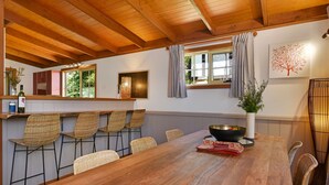 Un comedor con mesa de madera, sillas de mimbre, un amplio ventanal con cortinas y una obra de arte colgada en la pared.