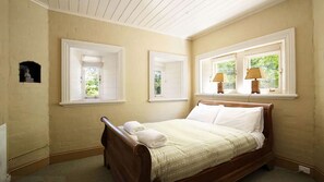 A bedroom with a wooden bed, two windows with white frames, and a small shelf with a statue.