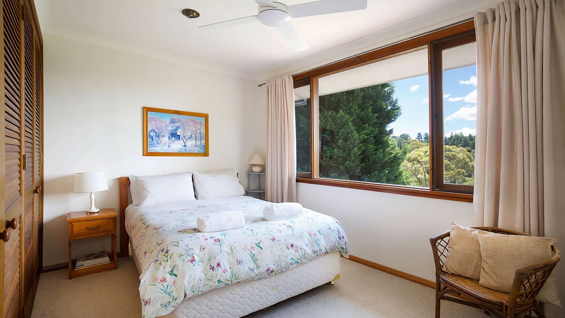 Une chambre à coucher comprenant un lit, une table de chevet, une chaise, une fenêtre avec des rideaux, un ventilateur de plafond et un tableau accroché au mur.