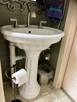 Ein weißes Waschtuch mit einem einzelnen Wasserhahn, einer Toilettenpapierrolle und einem Mülleimer in einem Badezimmer mit grünen Wänden und einem gemusterten Handtuch an der Wand.