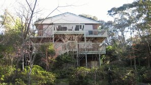 Une maison à deux étages avec un balcon au niveau supérieur, entourée d’une végétation luxuriante.