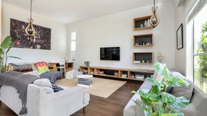 Una sala de estar moderna con un sofá, sillones, una mesa de centro y un televisor en la pared. Hay estanterías de madera con adornos y una planta grande en la esquina.