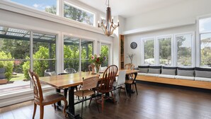 Un comedor espacioso con una mesa y sillas de madera, ventanas amplias que ofrecen vistas a zonas verdes y una araña.