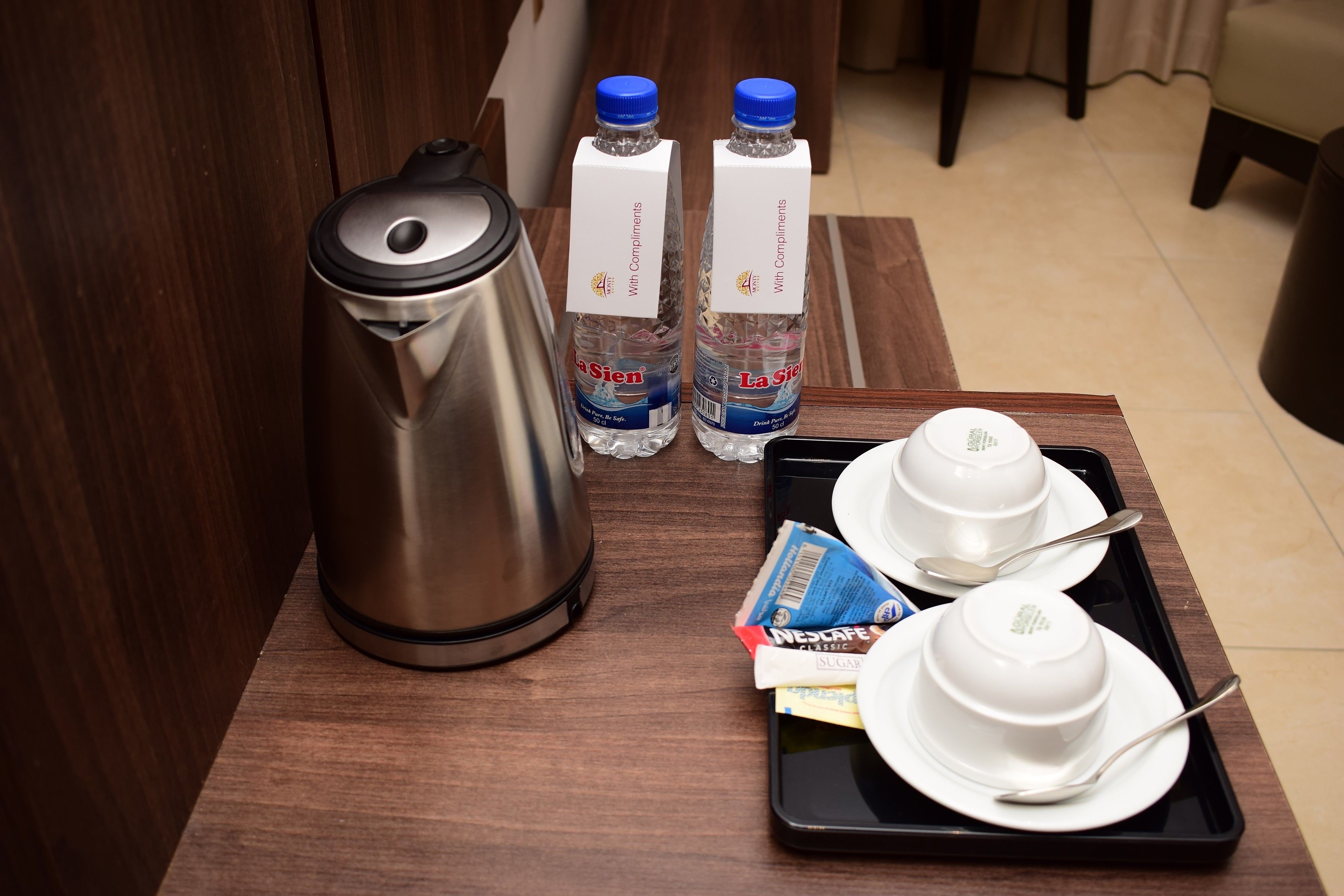 Habitación de hotel con una mesa de madera, un hervidor de acero inoxidable, dos botellas de agua, una bandeja con cuencos y cubiertos, y una caja de Nescaffe Classic Azúcar.