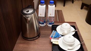 Habitación de hotel con una mesa de madera, un hervidor de acero inoxidable, dos botellas de agua, una bandeja con cuencos y cubiertos, y una caja de Nescaffe Classic Azúcar.
