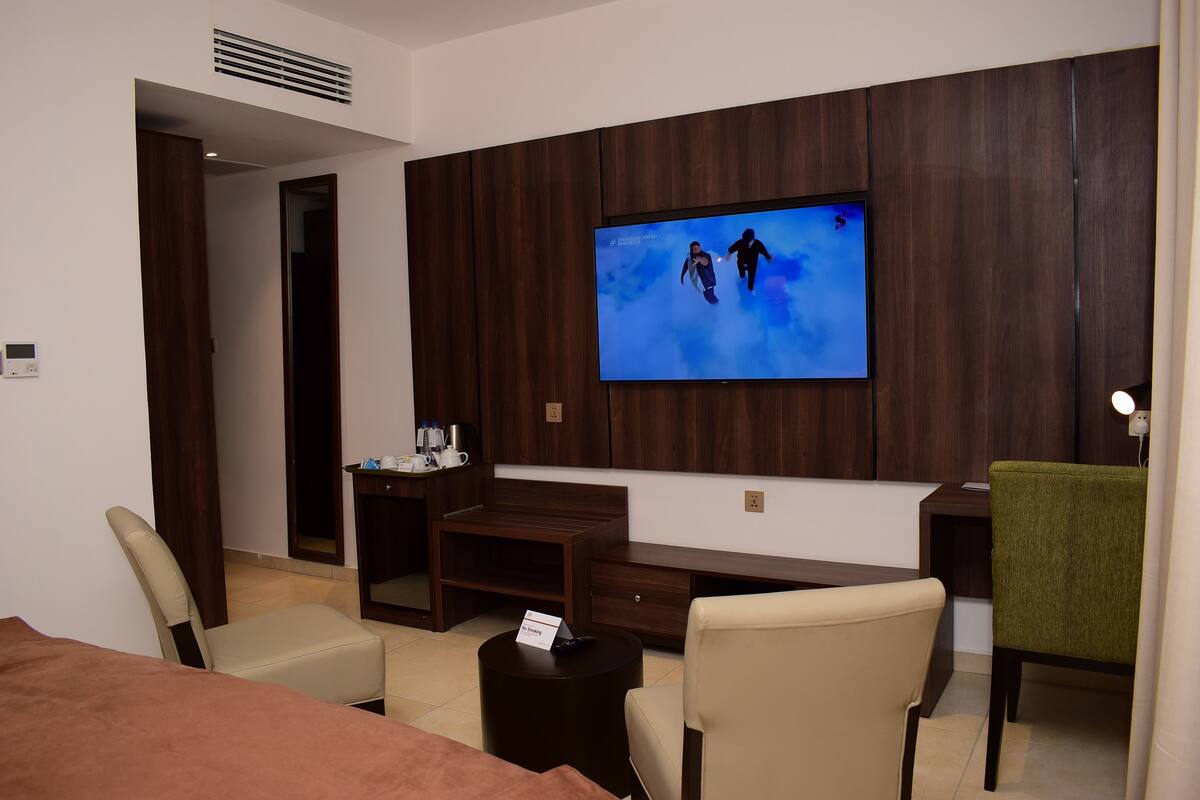 Quarto de hotel com televisão na parede, uma escrivaninha, uma cadeira, uma cama e uma mesinha com uma placa.