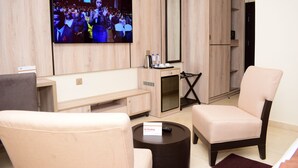 A modern hotel room with a flat-screen TV mounted on the wall, a sofa, a chair, a small coffee table, and a minibar. - Monty Suites & Golf (Uyo)