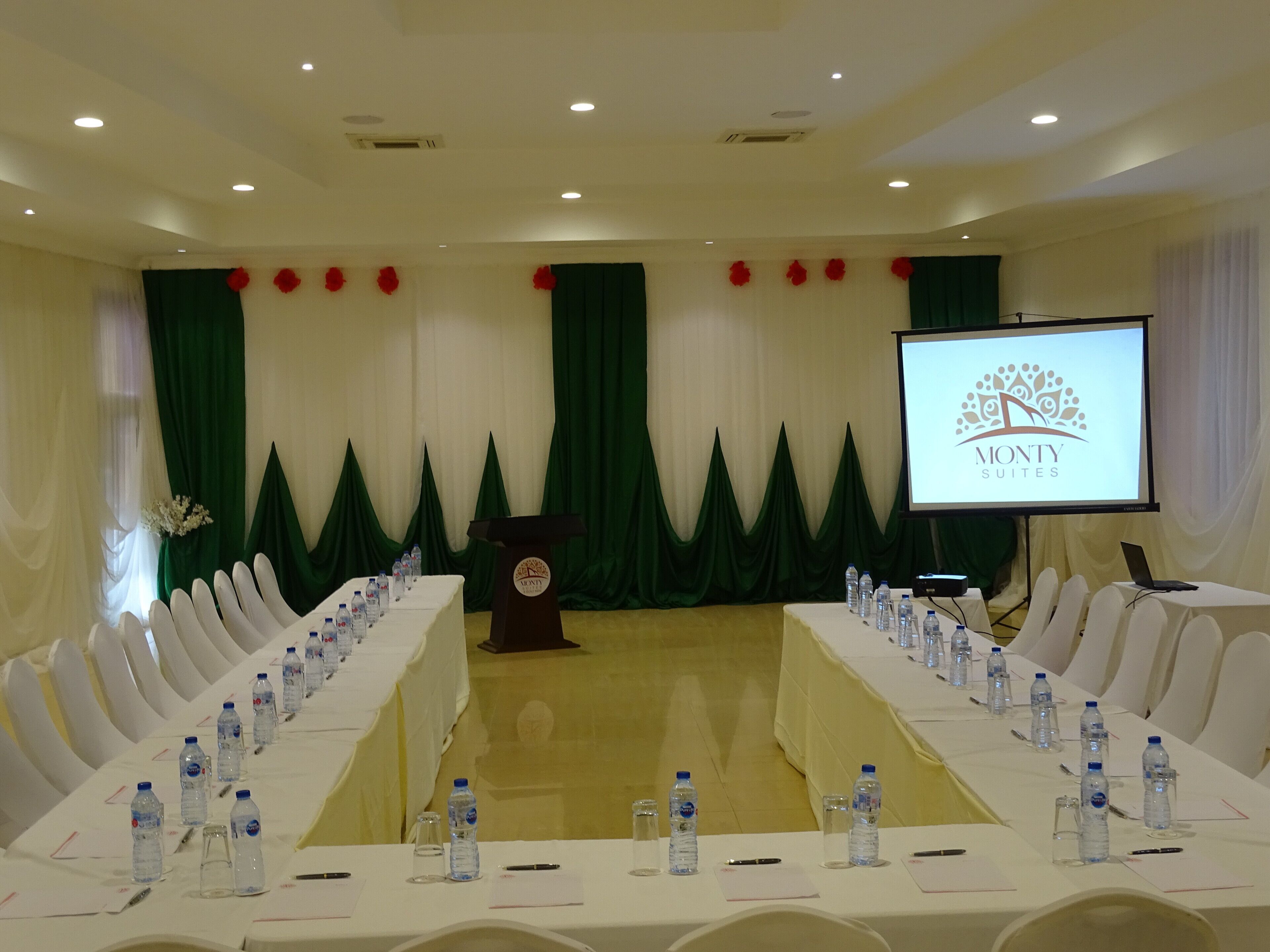 A conference room with a long table, chairs, water bottles, a podium, and a screen displaying a logo.