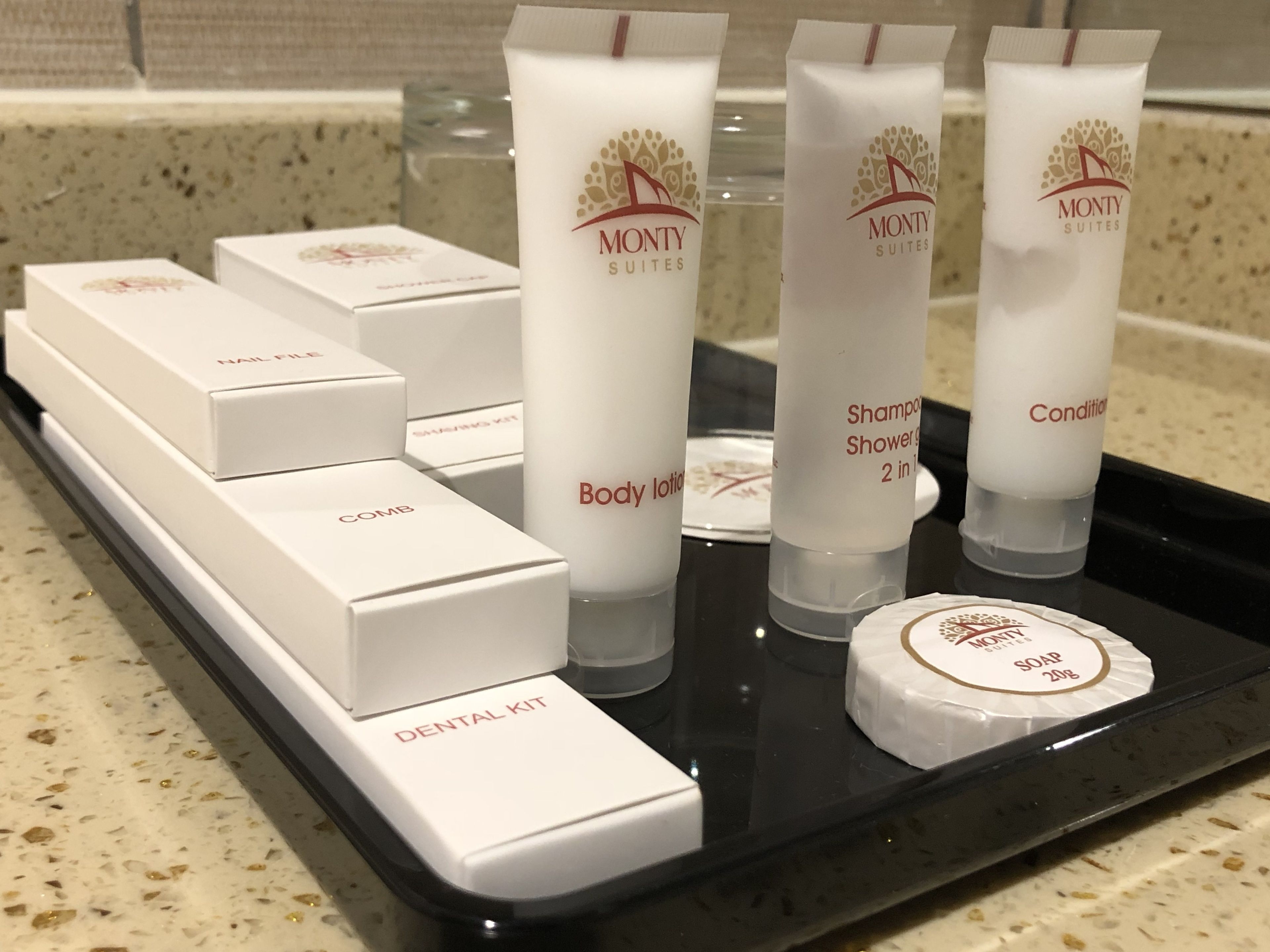 Een dienblad met toiletartikelen van Monty Suites, waaronder bodylotion, shampoo, conditioner en een tandverzorgingsset.